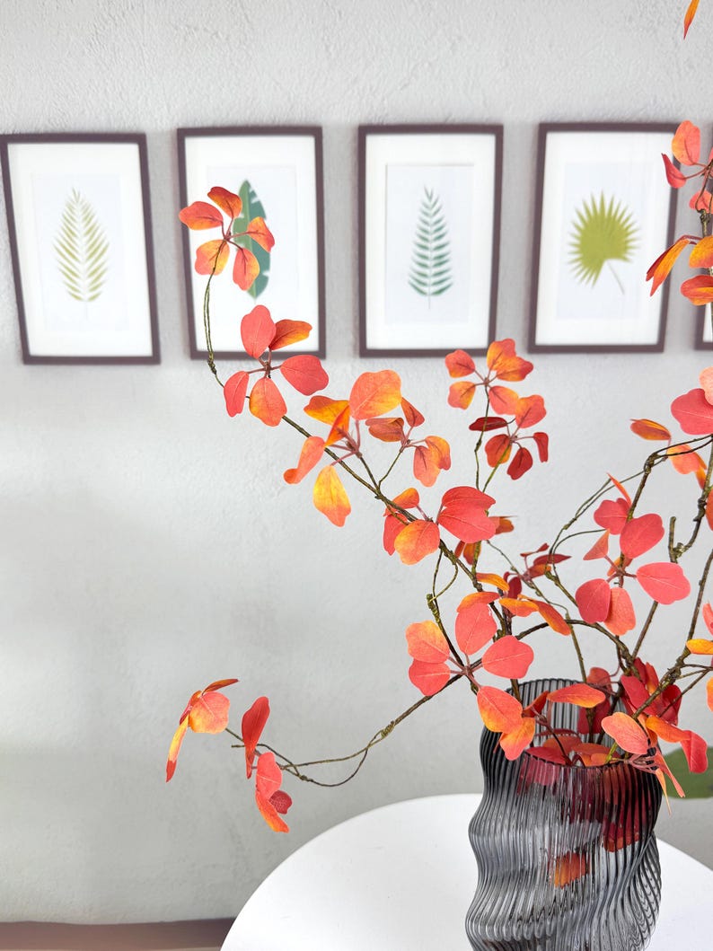 May include: A decorative arrangement featuring vibrant orange and red leaves in a textured gray vase. Framed botanical prints hang on the wall in the background, creating a harmonious display of nature-inspired decor.