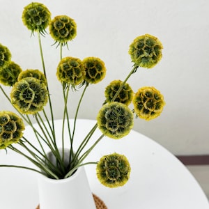 May include: A white ceramic vase with a narrow neck and a wide base, filled with a bouquet of dried yellow and green flowers. The vase is sitting on a woven brown coaster.