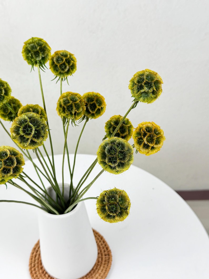 May include: A white ceramic vase with a narrow neck and a wide base, filled with a bouquet of dried yellow and green flowers. The vase is sitting on a woven brown coaster.