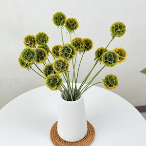 May include: A bouquet of dried green and yellow flowers in a white ceramic vase on a white table with a woven coaster.