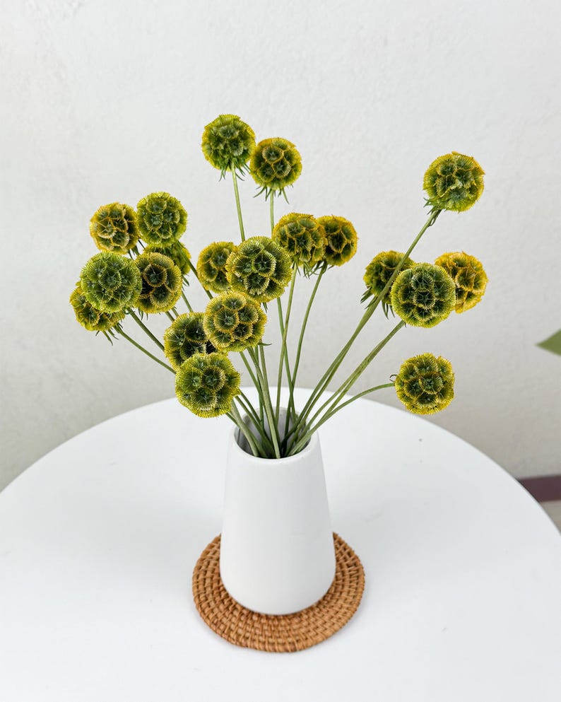 May include: A bouquet of dried green and yellow flowers in a white ceramic vase on a white table with a woven coaster.