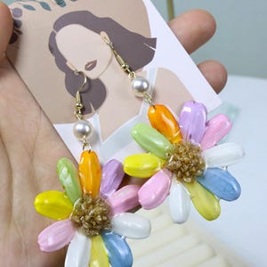 May include: A pair of colorful daisy earrings. Each earring features a gold-colored hook, a pearl, and a flower design with petals in various colors, including yellow, pink, blue, and orange. The center of each flower is a cluster of small, textured beads.