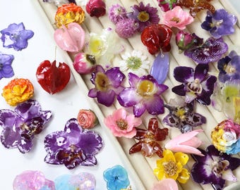May include: A collection of colorful resin flower pendants and earrings in various shapes and sizes. The assortment includes orchids, daisies, and other floral designs in shades of purple, pink, yellow, and red. Some pieces feature embedded glitter.