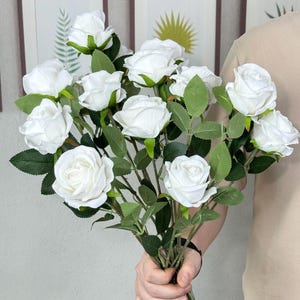 Puede incluir: Un ramo de rosas blancas con hojas y tallos verdes. Las rosas están en plena floración y las hojas son de un verde vibrante. El ramo se sostiene en la mano de una persona.