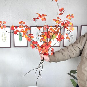 May include: A bouquet of artificial fall foliage with red and orange leaves. The stems are dark brown, and the leaves have a gradient of colors. The bouquet is held in front of a gray wall with framed botanical prints.