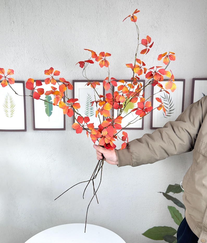 May include: A bouquet of artificial fall foliage with red and orange leaves. The stems are dark brown, and the leaves have a gradient of colors. The bouquet is held in front of a gray wall with framed botanical prints.