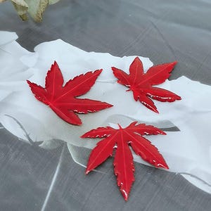 Può includere: Tre vivaci decorazioni a forma di foglia d'acero rossa con superfici lucide. Le foglie sono disposte su uno sfondo bianco e grigio, mettendo in risalto le loro venature e forma dettagliate. Queste foglie decorative sono ideali per lavori creativi a tema autunnale.
