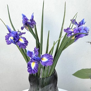 May include: A bouquet of purple iris flowers with yellow centers and green stems in a black, textured vase. The flowers are arranged in a vertical display, creating a visually appealing floral arrangement. The vase has a unique, wavy design.