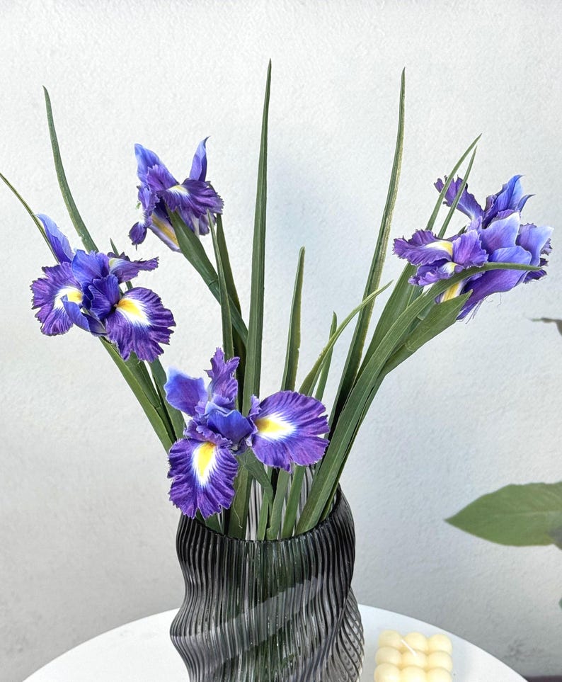 Puede incluir: Un ramo de flores de iris moradas con centros amarillos y tallos verdes en un jarr&oacute;n negro texturizado. Las flores est&aacute;n dispuestas verticalmente, creando un arreglo floral visualmente atractivo. El jarr&oacute;n tiene un dise&ntilde;o ondulado &uacute;nico.