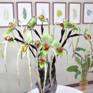 Puede incluir: Un arreglo decorativo de orquídeas artificiales en un jarrón negro. Las orquídeas tienen tallos verdes y marrones, con flores verdes y rojas. El jarrón está sobre una mesa redonda blanca. En el fondo se ven grabados botánicos enmarcados.