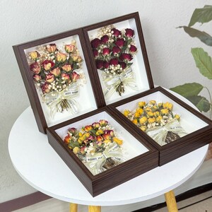Puede incluir: Cuatro arreglos florales enmarcados con rosas secas en tonos rojos, amarillos y crema, adornados con paniculata y cintas. Cada marco tiene un borde de madera marrón oscuro y un interior blanco.