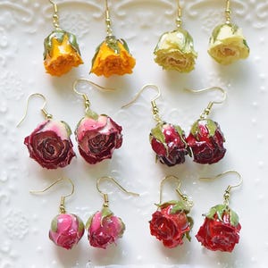 Op de afbeelding: Handgemaakte oorbellen met echte gedroogde rozen in verschillende kleuren, waaronder geel, roze en rood. Elke oorbel heeft een goudkleurige haak. De oorbellen worden gepresenteerd op een wit, decoratief oppervlak.