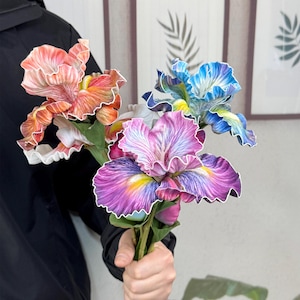 May include: A bouquet of four artificial iris flowers in shades of pink, blue, and orange. The flowers have a ruffled texture and are arranged in a hand.
