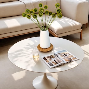 May include: A white ceramic vase with a round base sits on a woven coaster on a white round table. The vase holds a bouquet of green flowers with small, round blooms.