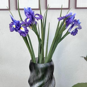 Puede incluir: Un ramo de flores de iris artificiales en un jarr&oacute;n de vidrio ondulado gris oscuro. Los iris tienen p&eacute;talos de color p&uacute;rpura intenso con detalles amarillos y blancos. El jarr&oacute;n est&aacute; sobre una superficie blanca, con hojas y tallos verdes.