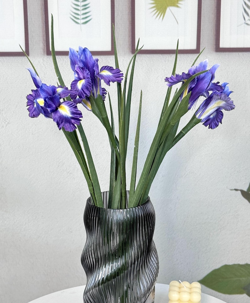 Puede incluir: Un ramo de flores de iris artificiales en un jarr&oacute;n de vidrio ondulado gris oscuro. Los iris tienen p&eacute;talos de color p&uacute;rpura intenso con detalles amarillos y blancos. El jarr&oacute;n est&aacute; sobre una superficie blanca, con hojas y tallos verdes.