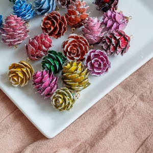 May include: A collection of colorful, miniature pine cone ornaments in various shades of red, pink, blue, green, and yellow. Each ornament has a gold-colored loop for hanging. The ornaments are arranged on a white rectangular plate.