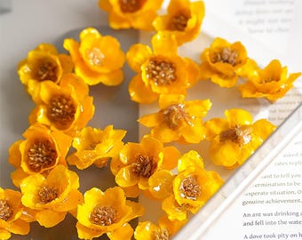 May include: A collection of bright yellow, resin-cast flowers with detailed brown centers. The flowers are scattered across a clear surface, creating a visually appealing arrangement. The petals are slightly curved, giving them a realistic appearance. The flowers are likely for crafting or decorative purposes.