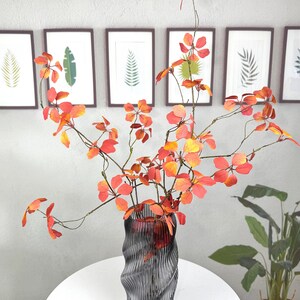 May include: A decorative arrangement featuring vibrant orange and red faux foliage in a gray glass vase. The vase has a unique, wavy design and sits on a round, woven coaster. Framed botanical prints hang in the background.
