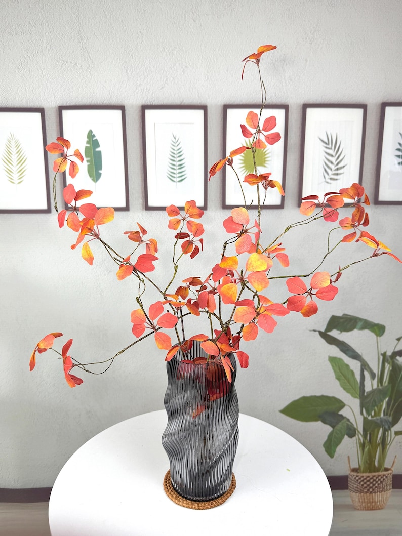 May include: A decorative arrangement featuring vibrant orange and red faux foliage in a gray glass vase. The vase has a unique, wavy design and sits on a round, woven coaster. Framed botanical prints hang in the background.