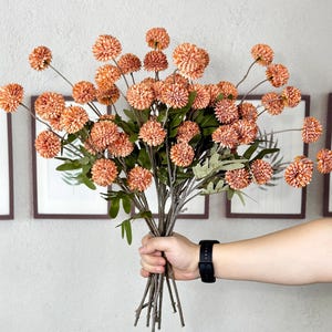 Puede incluir: Un ramo de flores artificiales naranjas con hojas verdes y tallos marrones. Las flores se sostienen en una mano que lleva un reloj negro. El fondo presenta obras de arte enmarcadas.