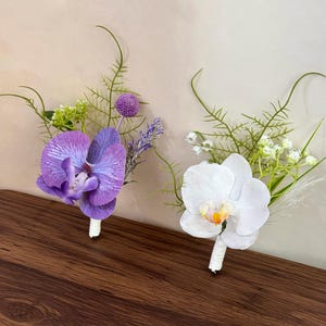 Op de afbeelding: Twee bloemenboutonnières met orchideeën. Eén corsage heeft een paarse orchidee met lavendelaccenten, de andere een witte orchidee. Beide hebben groen blad en zijn omwikkeld met wit lint.
