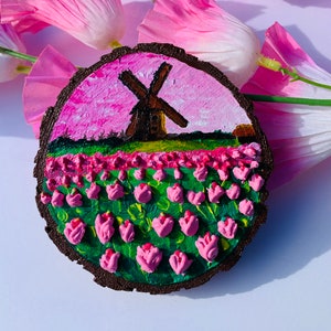 May include: A wooden round disc with a painted scene of a windmill in a field of pink tulips. The background is a pink and purple sky.