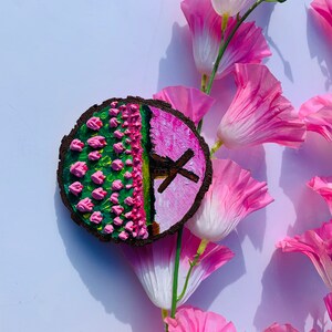 May include: A round wooden slice with a painted design of pink flowers and a windmill on a pink background. The slice is surrounded by pink flowers.