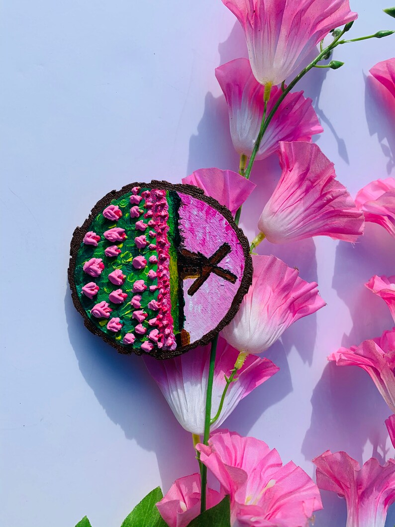 May include: A round wooden slice with a painted design of pink flowers and a windmill on a pink background. The slice is surrounded by pink flowers.