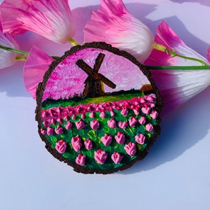 May include: A round wooden slice with a painted scene of a windmill in a field of pink tulips against a pink and white sky.