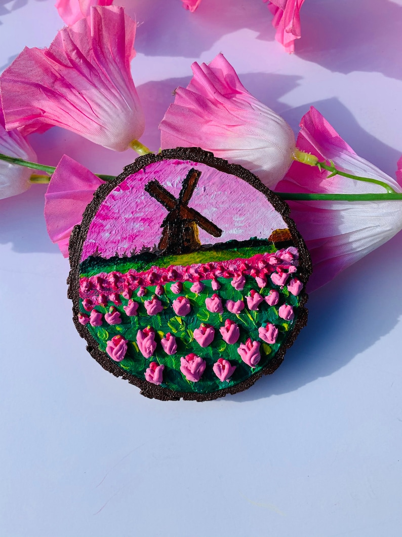 May include: A round wooden slice with a painted scene of a windmill in a field of pink tulips against a pink and white sky.