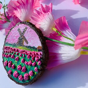 May include: A hand-painted wooden round ornament with a pink and green landscape featuring a windmill and a field of pink tulips.