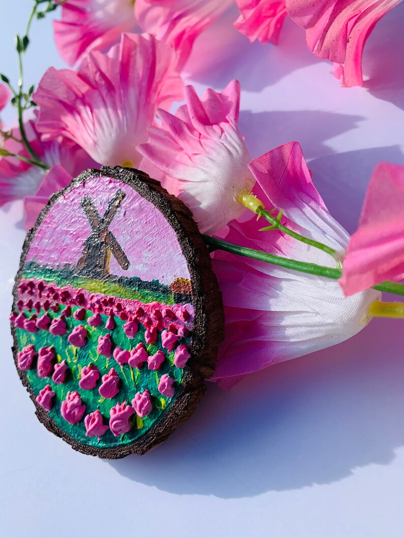 May include: A hand-painted wooden round ornament with a pink and green landscape featuring a windmill and a field of pink tulips.