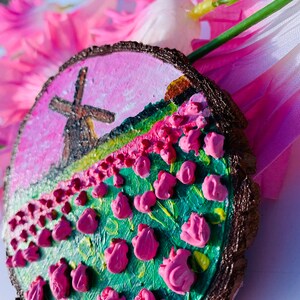 May include: A hand-painted wooden slice with a pink and green landscape featuring a windmill and pink flowers. The slice is painted with a textured, 3D effect.