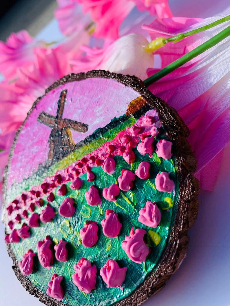 May include: A hand-painted wooden slice with a pink and green landscape featuring a windmill and pink flowers. The slice is painted with a textured, 3D effect.