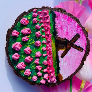 May include: A round wooden slice painted with a pink and green landscape featuring a windmill and pink flowers. The background is a light pink color.
