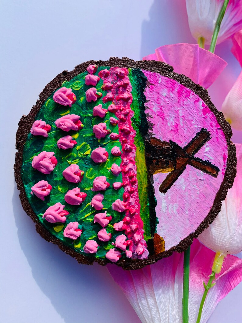 May include: A round wooden slice painted with a pink and green landscape featuring a windmill and pink flowers. The background is a light pink color.