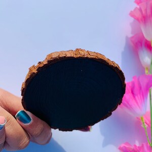 May include: A round wooden coaster with a dark brown bark edge and a black painted surface. The coaster is being held by a hand with blue nail polish.