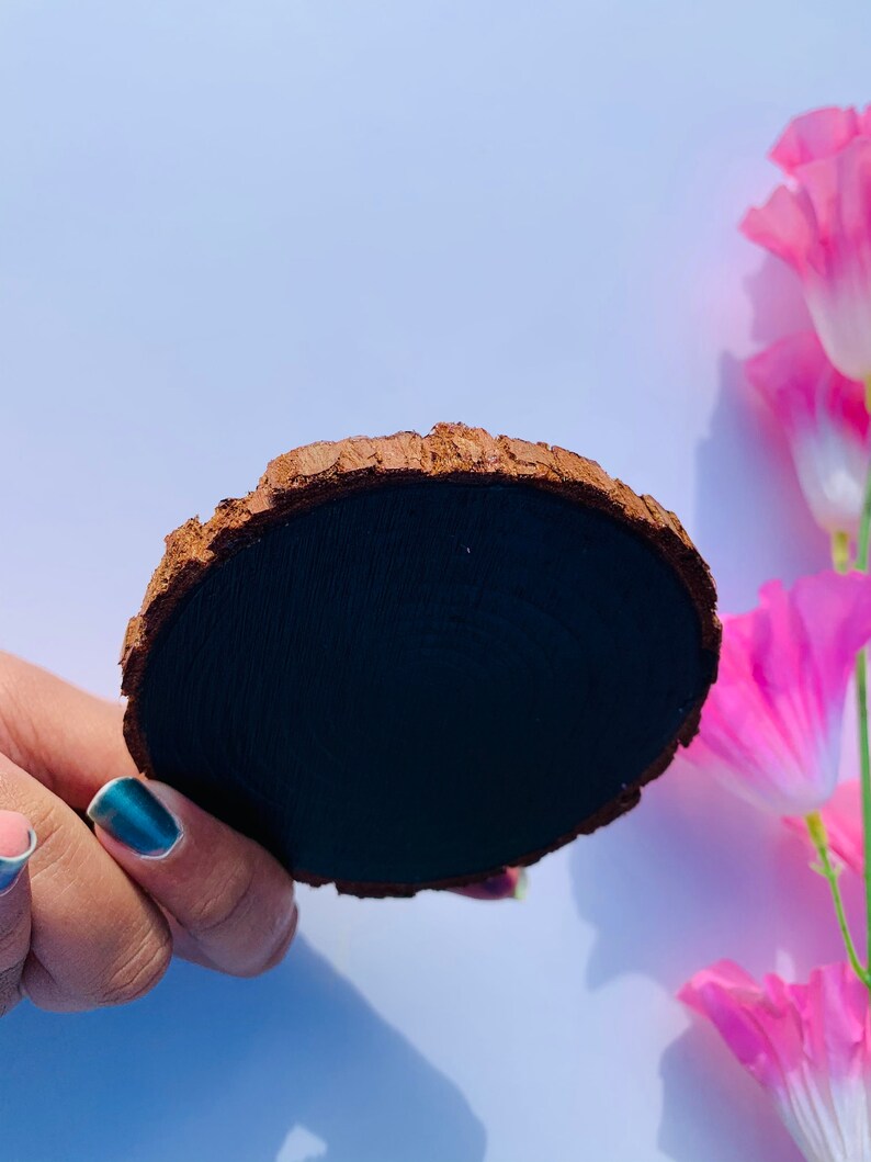May include: A round wooden coaster with a dark brown bark edge and a black painted surface. The coaster is being held by a hand with blue nail polish.