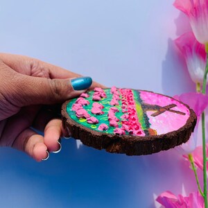 May include: A round wooden slice with a painted scene of a pink field with a cross in the background. The field is painted with green and yellow accents. The wood is brown and has a natural bark edge.