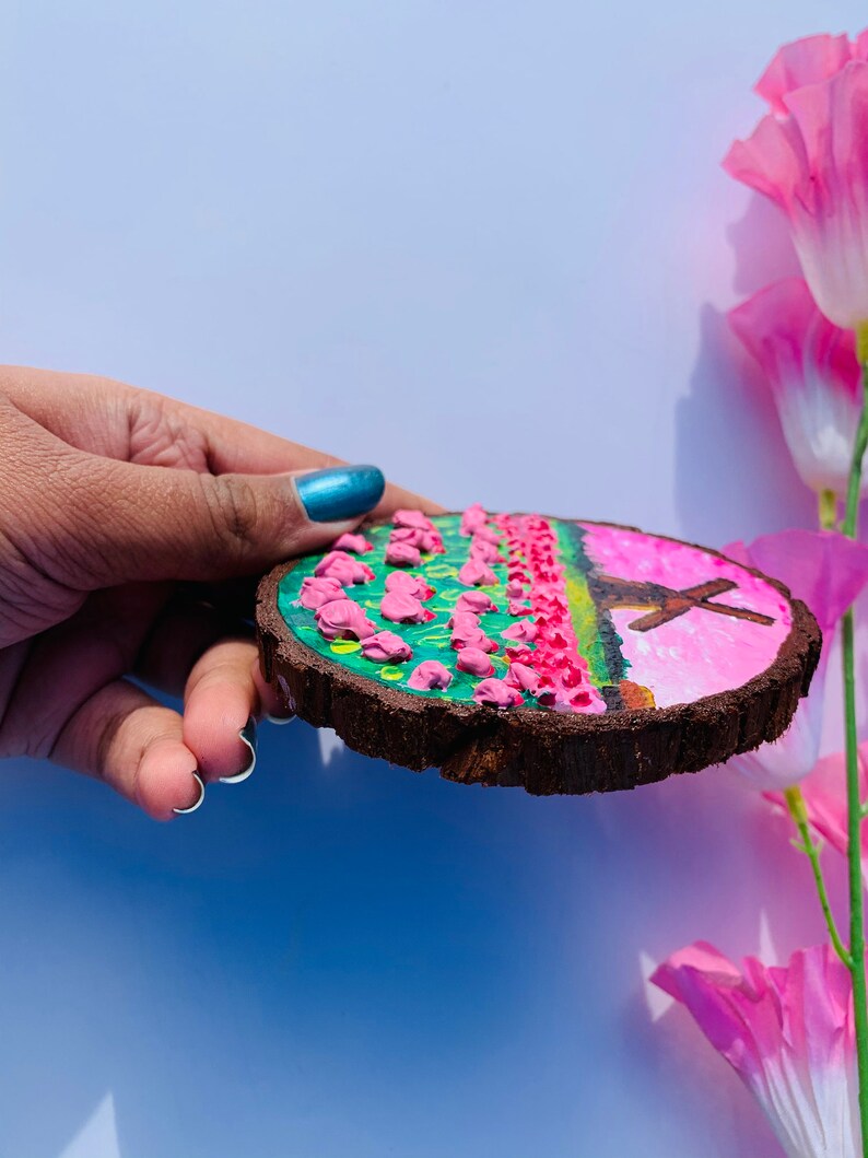May include: A round wooden slice with a painted scene of a pink field with a cross in the background. The field is painted with green and yellow accents. The wood is brown and has a natural bark edge.