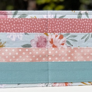 May include: A rectangular fabric organizer with multiple horizontal pockets. The pockets feature various floral and polka dot patterns in shades of pink, blue, and teal. The edges are neatly stitched, creating a decorative and functional storage item.