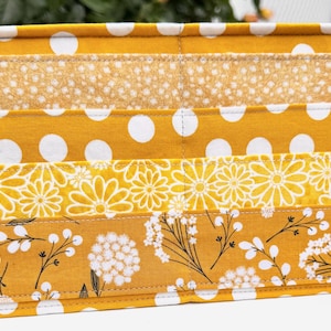 May include: A rectangular fabric organizer with a patchwork design. The design features horizontal bands of yellow fabric with white polka dots, floral patterns, and a dotted texture. The edges are finished with white stitching.