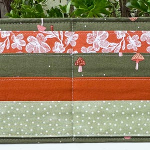 May include: A fabric organizer with multiple pockets. The design features horizontal stripes in various colors and patterns, including floral prints, solid orange, and polka dots. Small mushroom appliqués are scattered across the surface.