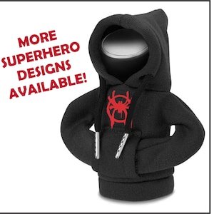 May include: A black hoodie-style can cooler with a red spider emblem on the front. The cooler has a drawstring and metal aglets. The text "MORE SUPERHERO DESIGNS AVAILABLE!" is in red on the left side. The cooler is designed to keep beverages cold.