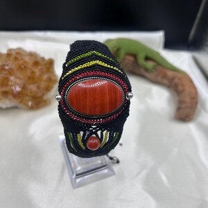 Peut inclure: Un bracelet en macramé noir orné d'une grande pierre ovale brun-rouge sertie dans un cadre argenté. Le tissage intègre des fils fins jaunes et rouges, agrémenté d'une petite perle ronde brun-rouge située sur la partie inférieure.