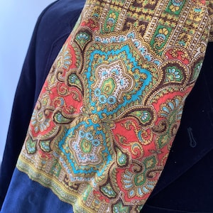 Puede incluir: Una bufanda con estampado paisley con un diseño rojo, azul, verde y dorado. La bufanda está doblada y drapeada sobre una chaqueta azul marino.