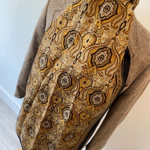 May include: A paisley patterned scarf in shades of gold, brown, and black. The scarf is draped over a brown blazer. The intricate design features floral and geometric elements. The scarf is made of a silky material.