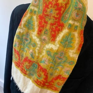 Op de afbeelding: Een witte sjaal met een kleurrijk paisley-patroon in tinten geel, rood en groen. De sjaal heeft een gefranst randje.