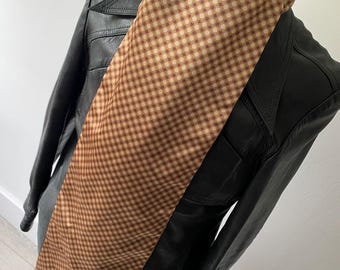 May include: A brown and tan checkered scarf with black fringe is draped over a black leather jacket on a mannequin. The scarf has a repeating pattern of small squares in shades of brown and tan. The jacket has a classic design.